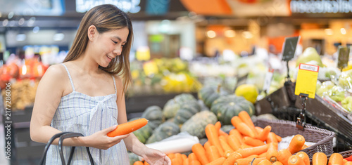 Happy asian woman holding fresh carrot in supermarket produce section, Smiling female customer buying healthy nutritious foods, Vegan vegetarian lifestyle and modern retail grocery shopping concepts