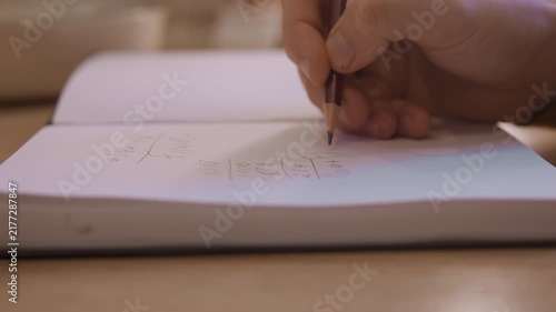 Close up of hand writing mathematical equations in pencil on white notebook paper showing detailed arithmetic calculations. scene features soft lighting and natural indoor setting