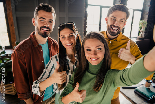 Fotografie Four happy coworkers taking a selfie with thumbs up in a modern office, celebrat