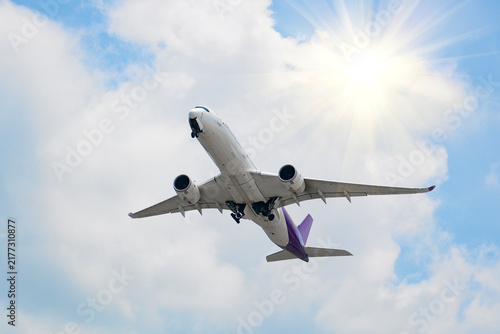 Modern jet airplane soaring high into the atmosphere with landing gear retracting against a backdrop of soft white clouds on a sunny day