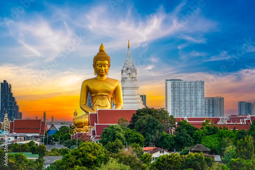 Large golden Buddha sits peacefully next to a white pagoda surrounded by lush trees and city buildings under a vibrant sunset sky in Bangkok