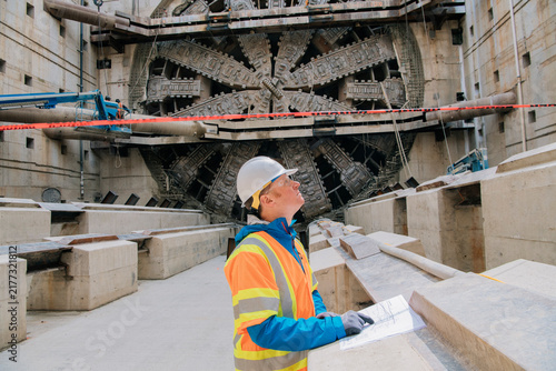 A construction white worker inspects blueprints by a tunnel boring