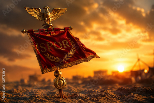 A red Roman legionary standard with a golden eagle planted in sandy soil.