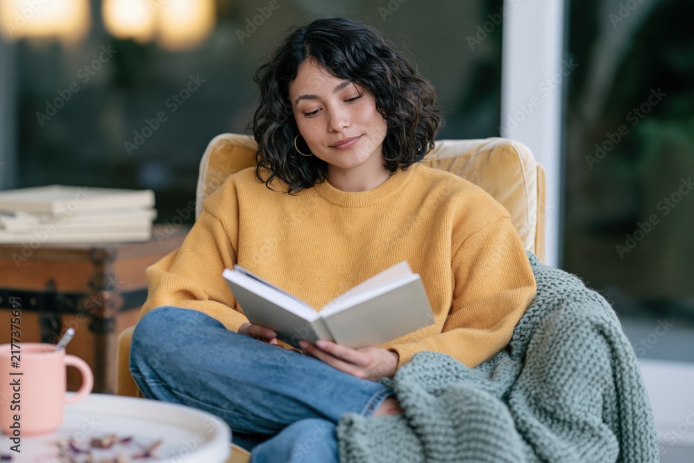 Obraz premium Closeup portrait of a young woman reading a book in a yellow sweater on a knitted blanket for calm lifestyle and relaxation wallpaper