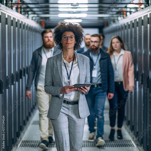A diverse group of professionals walking through a modern data center, led by a confident woman holding a tablet