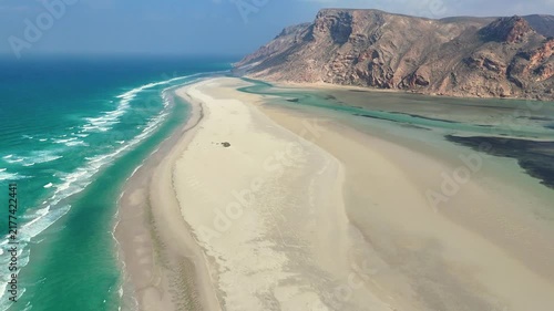 Socotra island, Aerial view of a vast sandy coastline where desert dunes and rugged mountains meet turquoise ocean lagoon waters, with dramatic shadows and bright sunlight reflecting across the sea.