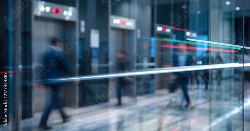 A busy lobby area showcasing blurred figures walking past glass elevators, conveying a sense of motion and modern architecture in an urban environment.