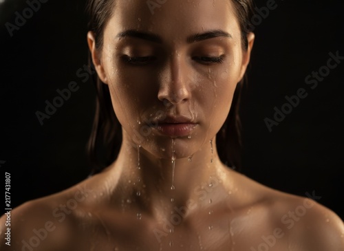 Close-up of a young woman with closed eyes and water dripping down her face and neck on a dark background, concept of purity and freshness
