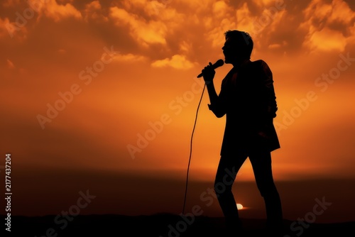 Plakat Performer silhouette holding a microphone and singing on an outdoor stage at sunset