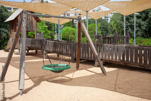 Basket swing and wooden playground equipment located in a public city park under shade sails surrounded by green trees. Outdoor recreational space designed for children, families.
