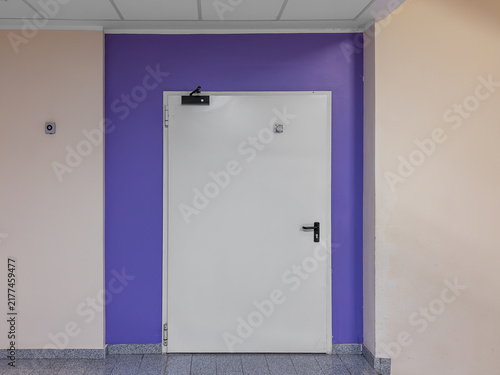 Single leaf white metal fire exit door with black handle and automatic closer hydraulic system installed on a purple painted wall section in a modern building hallway or public office or hospital