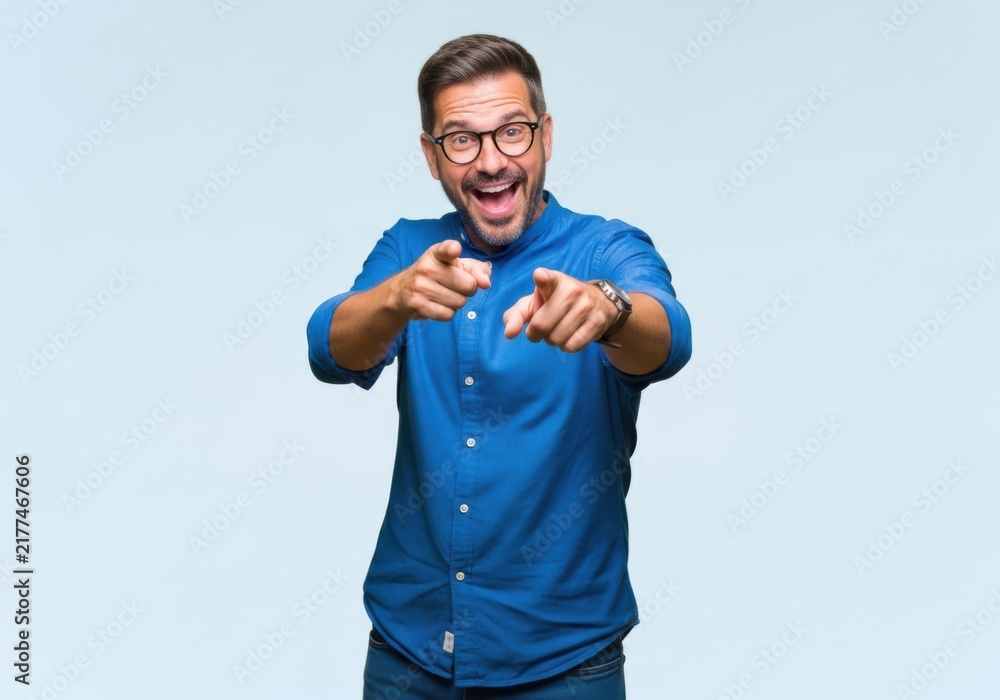 Fototapeta premium Excited middleaged man with glasses wearing a blue shirt pointing directly at the camera with both hands, expressing joy and enthusiasm against a light background