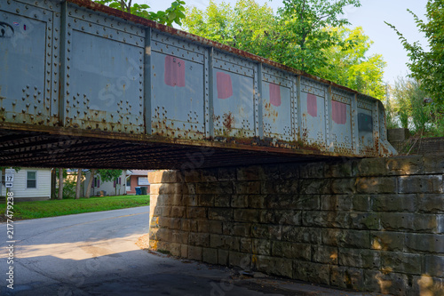 Принти на полотні Rusted metal rail bridge spans a narrow town road above a weathered stone abutment, framed by green trees and suburban houses, evoking a quiet, nostalgic and industrial small-town atmosphere