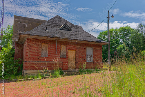 Quadro su tela An abandoned Pennsylvania Railroad red brick train station with boarded windows, an overgrown platform and adjacent railroad tracks beneath a bright blue sky