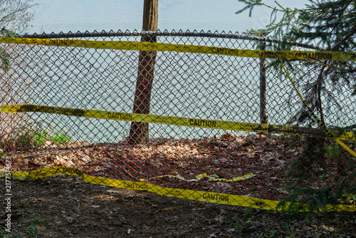 Yellow caution tape surrounds a chain link fence and wooded ground, creating a restricted and abandoned zone