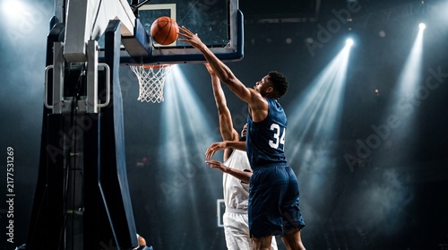 Basketball players compete for the ball under dramatic stadium lighting.