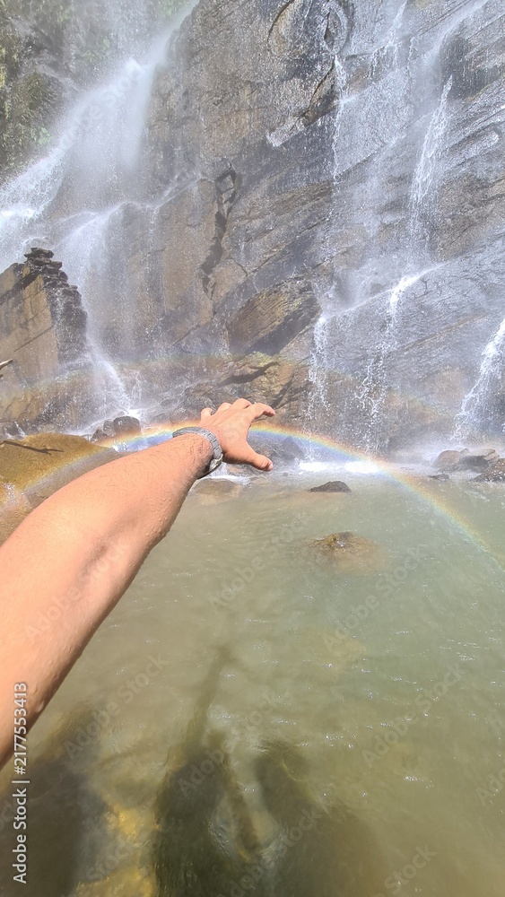 Obraz premium A person's hand reaches out to touch a beautiful rainbow forming in the mist of a powerful waterfall cascading down rocky cliffs into a clear pool of water