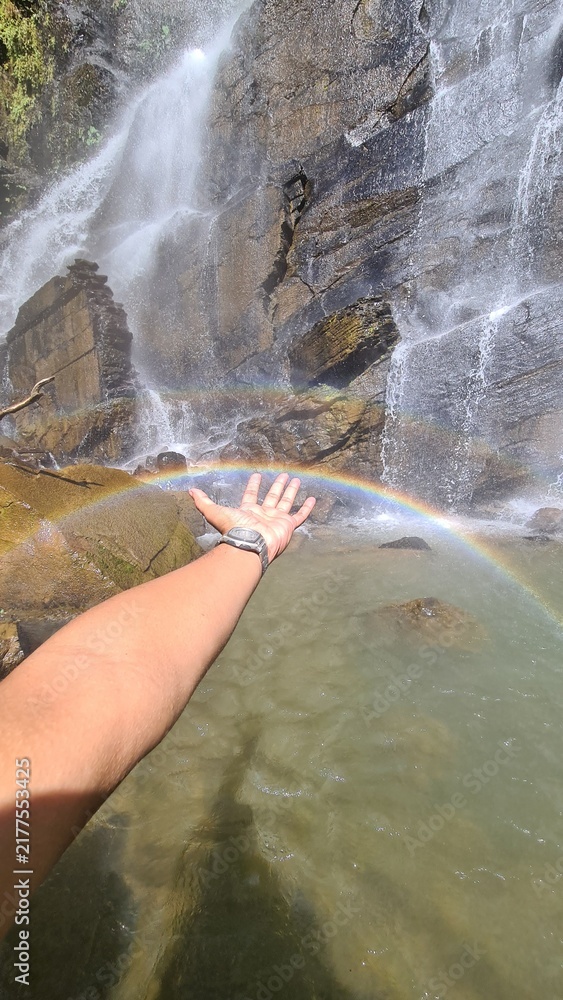 Obraz premium A hand reaching towards the vibrant spectrum of a rainbow, gracefully arcing over the tranquil waters at the base of a powerful waterfall, a serene scene of natural beauty and wonder