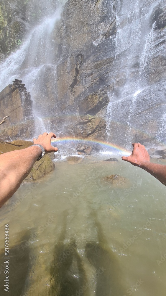 Obraz premium Person holding a vibrant rainbow between their hands in front of a majestic mountain waterfall cascading into a clear pool