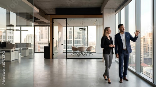 Professional businessman and businesswoman in formal attire walking through a modern, spacious office with large windows overlooking a sprawling urban city skyline at daytime.
