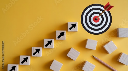 Wooden blocks with arrows pointing towards a target with a dart on a yellow background, for business achievement