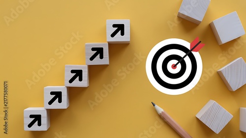 Wooden blocks with arrows pointing up towards a target with a dart on a yellow background, for business strategy
