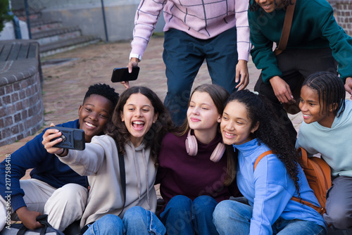 Diverse friends with backpacks, seven teens and one adult posing, holding smartphone on courtyard