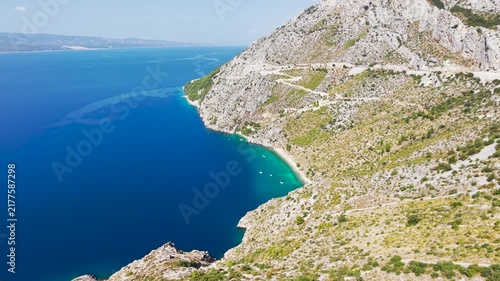 Aerial drone view of Vruja beach a secluded Adriatic bay with crystal-clear turquoise water, rocky cliffs, a coastal road and small boats along the Croatian Dalmatian coast.
