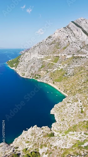Aerial drone view of Vruja beach a secluded Adriatic bay with crystal-clear turquoise water, rocky cliffs, a coastal road and small boats along the Croatian Dalmatian coast.