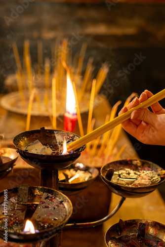 Fotografía An intimate view of a cultural tradition, showing a devotee's hand carefully dipping yellow joss sticks into a burning oil basin to light them for prayer inside a historic temple