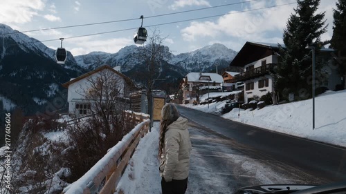 Woman in white coat stands on snowy road. She gazes at distant mountains. Ski lifts glide over snow-covered peaks. Snowy village with chalets fills background. Calm, contemplative mood