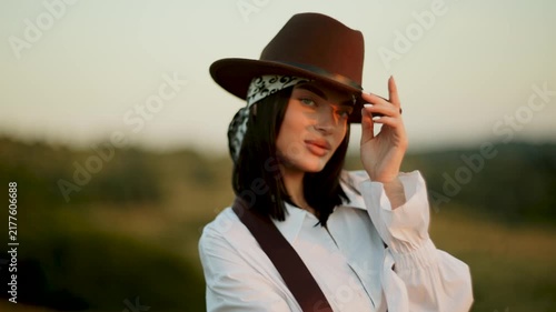Young woman wearing a brown hat and scarf poses outdoors during a golden sunset in the countryside