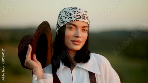 Woman wearing a brown hat and white shirt standing in a grassy field during a warm golden sunset
