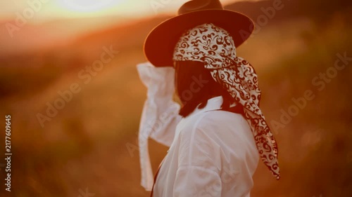 Young woman wearing a patterned headscarf walks through a grassy meadow during a warm golden sunset