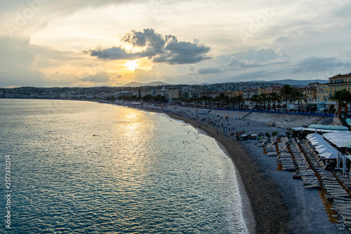 Sunset over Nice, France with calm Mediterranean waters, curved shoreline, beach loungers, palm‑lined promenade, and warm golden light creating a serene coastal atmosphere.