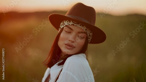Stylish woman wearing a brown hat looks away in a grassy field during a beautiful golden sunset