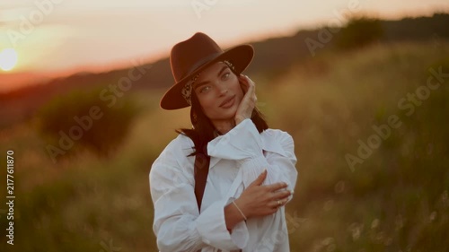 Woman wearing a stylish hat poses in a meadow during a golden sunset in the countryside