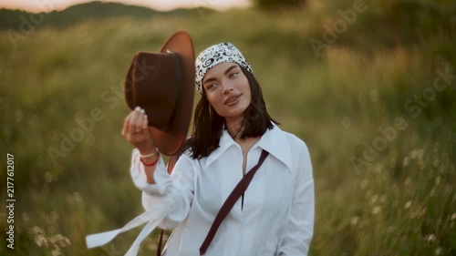 Young woman posing in a brown hat and white shirt within a grassy field during the golden hour