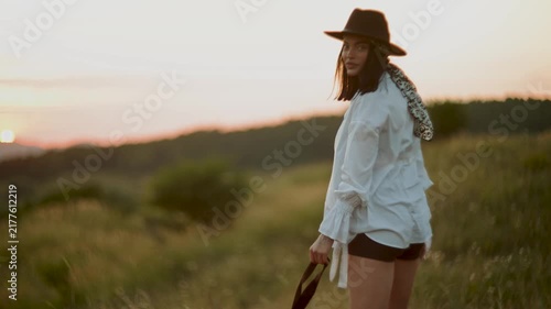 Woman in a wide-brim hat walking through a grassy field at sunset