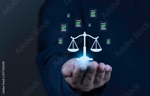 Compliance with laws and regulations concept with businessman hand holding glowing virtual scales of justice and verified document icons with green checkmarks on dark background.