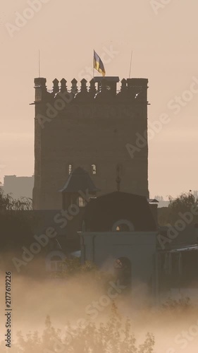 Vertical video. Silhouetted medieval castle tower with the ukrainian flag waving against a hazy sky as thick morning fog rolls through the city, creating a mystical and historical atmosphere
