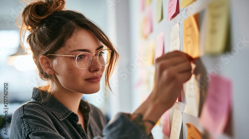 Planning and Organization: A focused woman with her hair in a bun, wearing glasses, is attaching sticky notes to a wall covered with them. This image represents planning, organization.