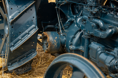 Foto Vintage blue tractor detail with steel lug wheel, engine block, and belt drive pulley standing on stubble agricultural field during harvest exhibition