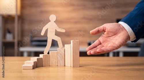 Experienced hand guides a wooden figure up a staircase of blocks symbolizing career growth and mentorship