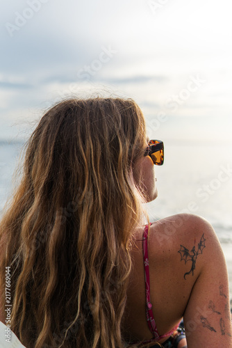 Canvastavla Woman with long wavy hair and sunglasses gazes at calm ocean from sunlit beach,