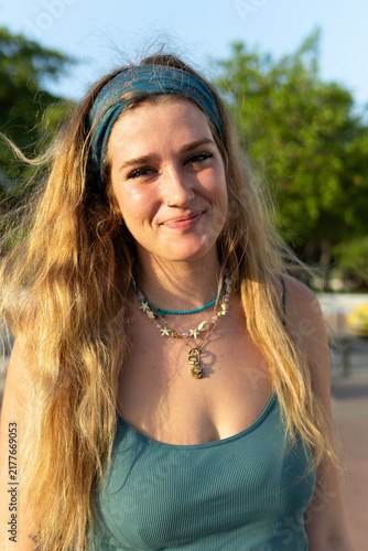 Young blonde woman smiling, wearing a headband and layered necklaces, enjoying golden hour sunlight outdoors, portraying natural beauty, freedom, and summer vibes