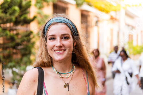 Young woman smiling at camera, wearing a headband and necklaces, expressing joy and confidence while traveling through a vibrant city street under warm sunlight