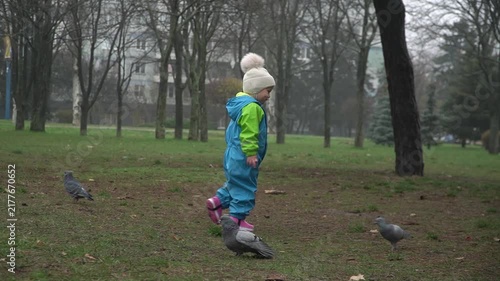 A girl in a blue and green jumpsuit slowly walks among a flock of pigeons, scaring them, a rainy autumn day, coolness, the joy of a child