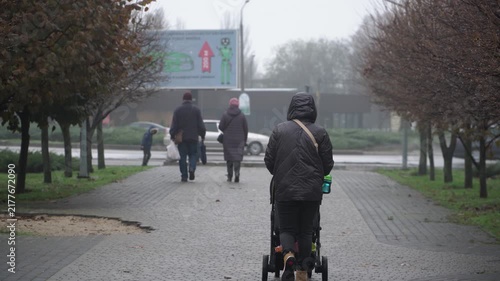 single mother, raising child alone. black jacket jeans hood over head pushes stroller sleeping autumn rain city park. cool day, filmed from side. trunk stroller full toys. background busy road with
