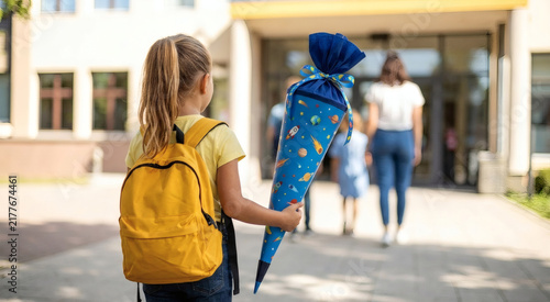 Fototapeta kuchenna Mädchen von hinten mit gelbem Schulrucksack und großer blauer Schultüte vor Grundschule, Erwachsene und Kind am Schuleingang